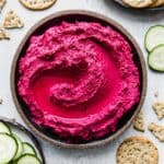 Beet Hummus on a round black plate surrounded by sliced cucumbers and round whole grain crackers.