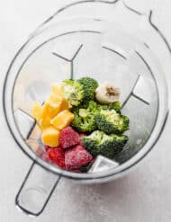 A vitamix blender full of raw broccoli, strawberries, mango, and frozen banana in preparation to make a broccoli smoothie.