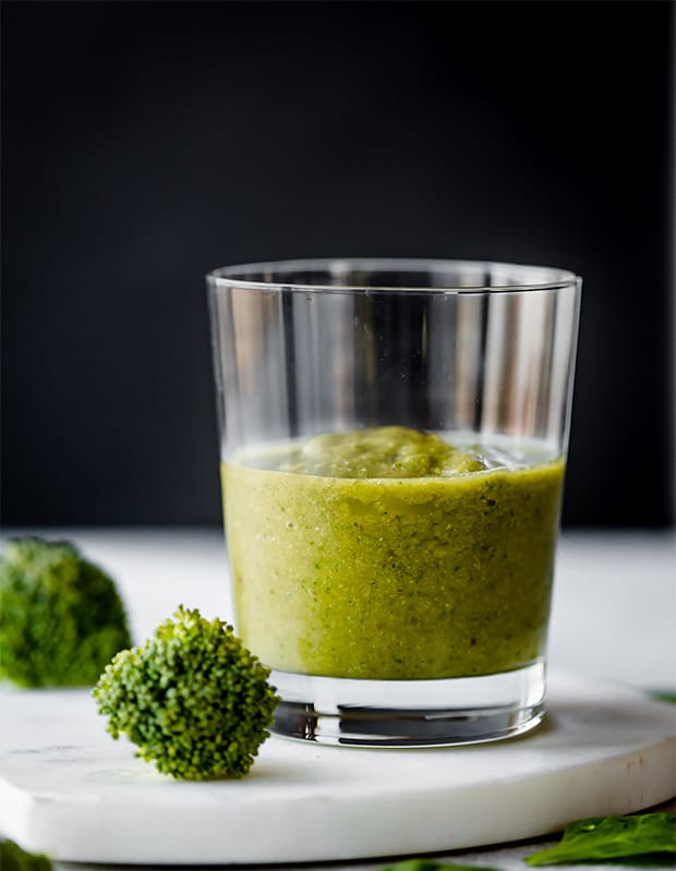 A green broccoli smoothie in a glass cup surrounded by 2 broccoli florets.