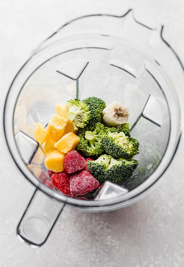 En vitamixblender full av rå broccoli, jordgubbar, mango och fryst banan som förberedelse för att göra en broccolismoothie.