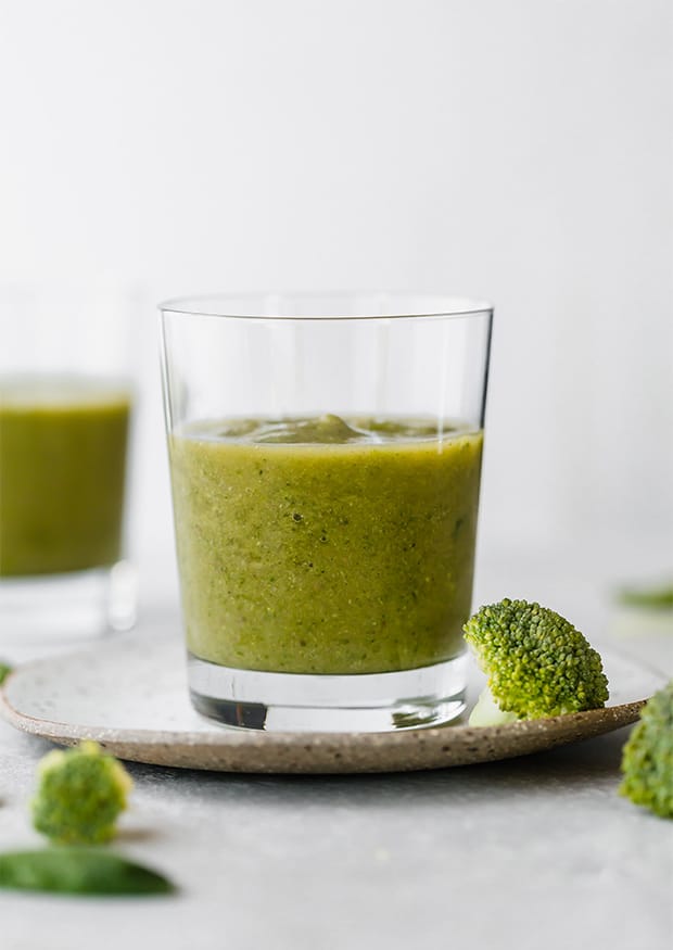 A green broccoli smoothie in a glass cup with some broccoli florets scattered around the cup.