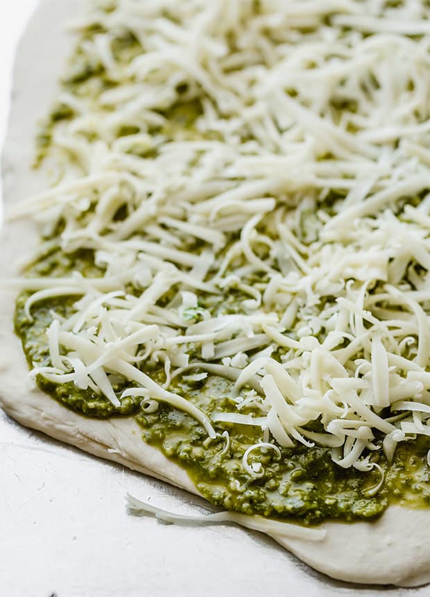 Uncooked pizza dough with basil pesto and shredded mozzarella.
