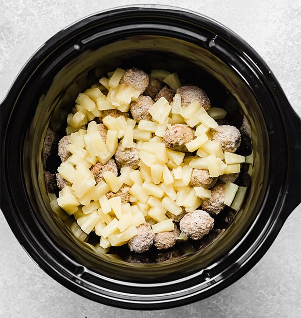 A black slow cooker with frozen meatballs and pineapple chunks in it. 