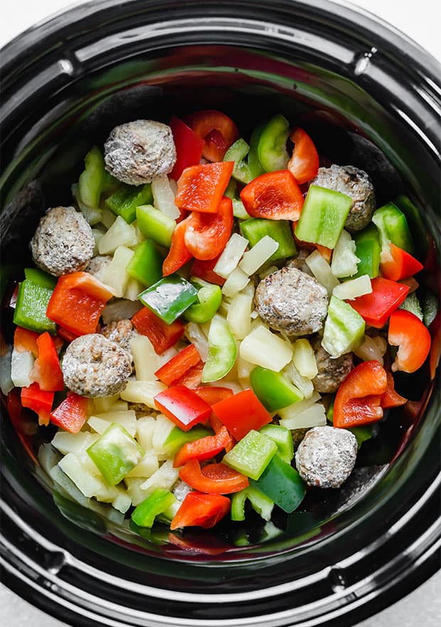 A crock pot with frozen meatballs, red and green chopped bell peppers, and pineapple tidbits.