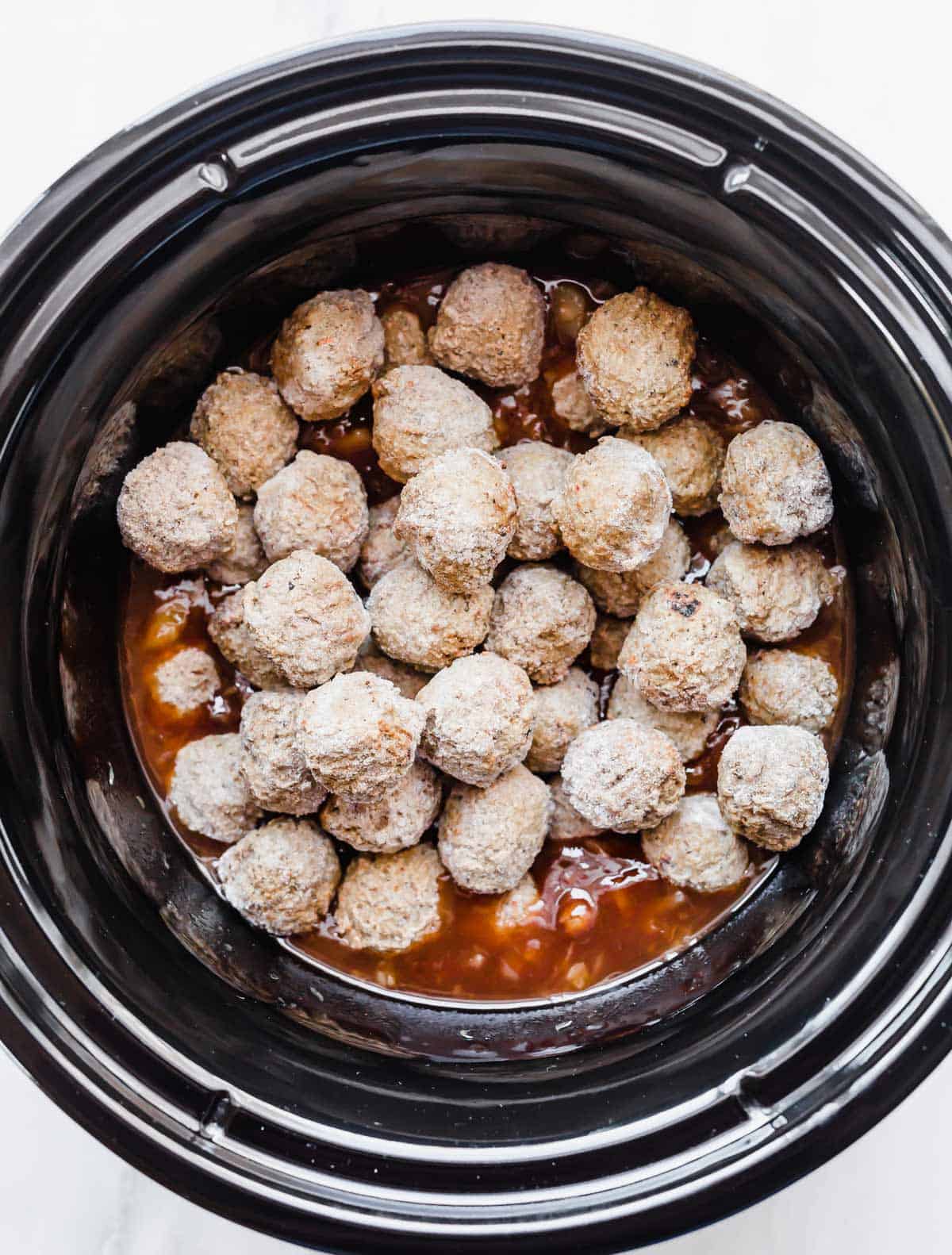 A black crockpot filled with frozen meatballs atop BBQ sauce and crushed pineapple.