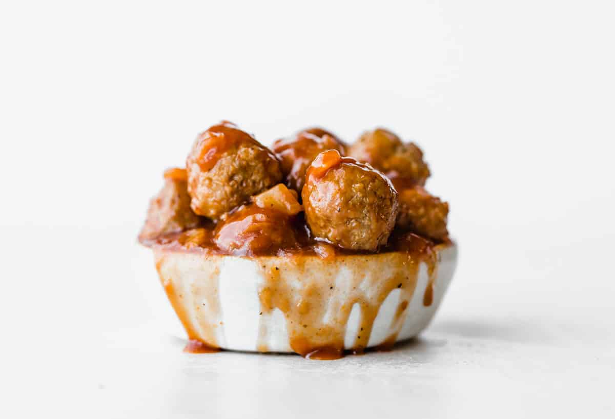 BBQ Pineapple Meatballs and sauce in a bowl against a white background with BBQ sauce spilling over the bowl.