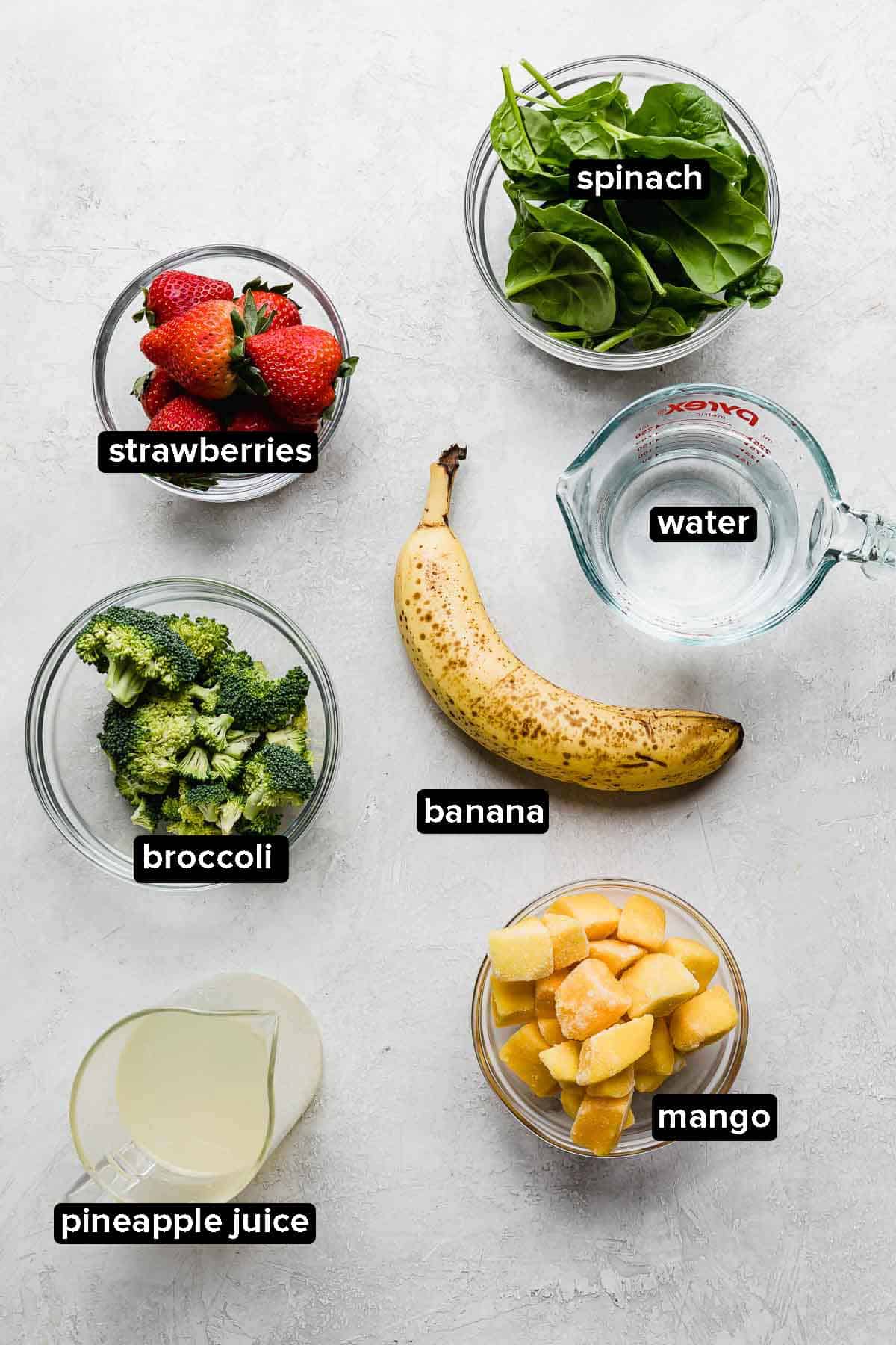 Broccoli Smoothie ingredients on a white background: strawberries, spinach, broccoli, banana, mango, pineapple juice, and water.