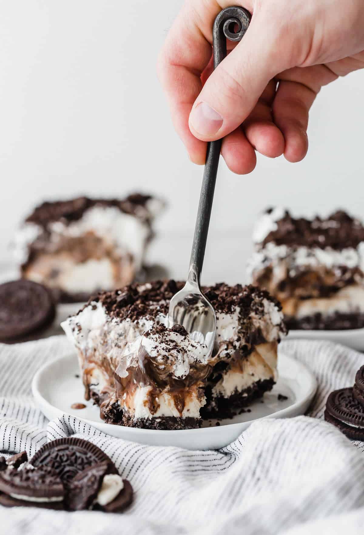A hand stabbing a fork into the front of an Oreo Ice Cream Cake slice, on a white plate.