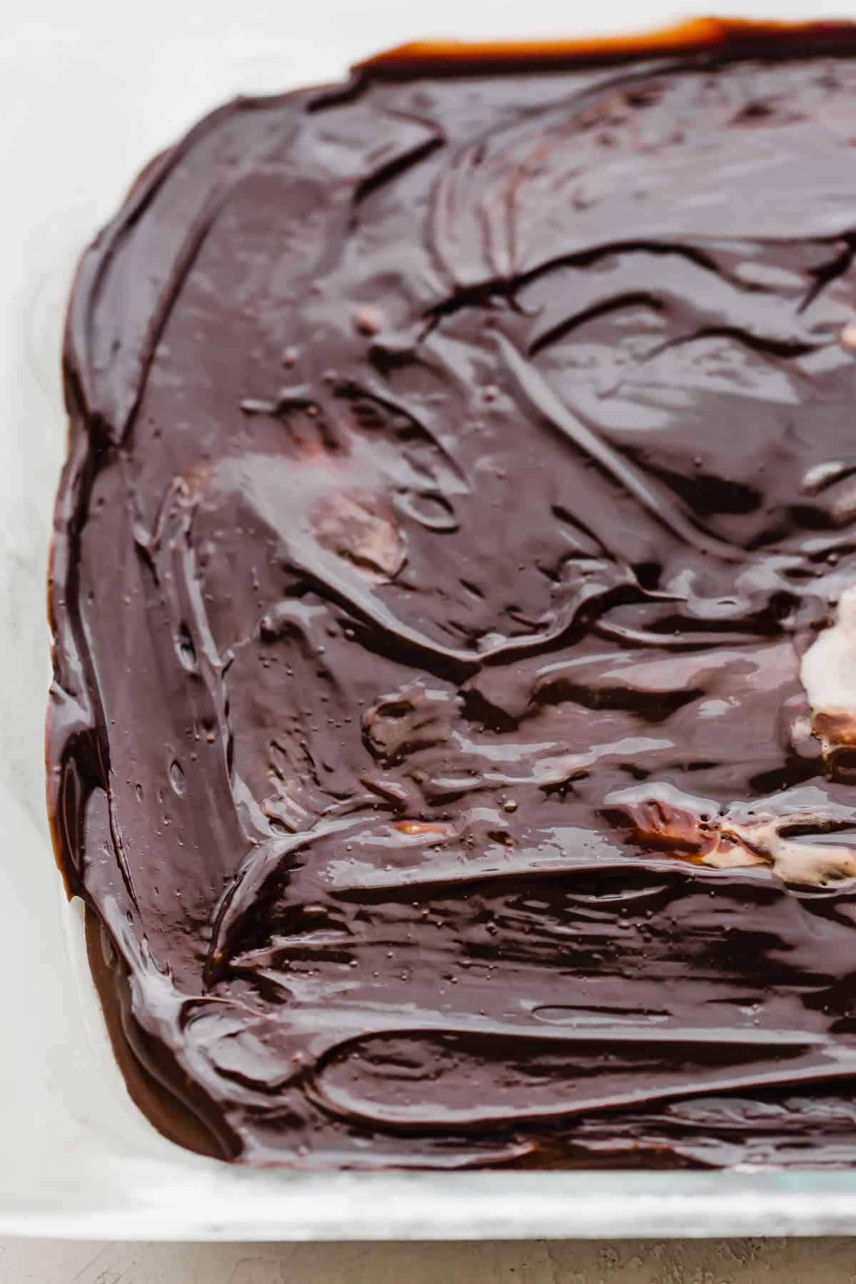 A close up photo of hot fudge overtop Oreo ice cream that has been spread into a baking dish, for making Oreo Ice Cream Cake.
