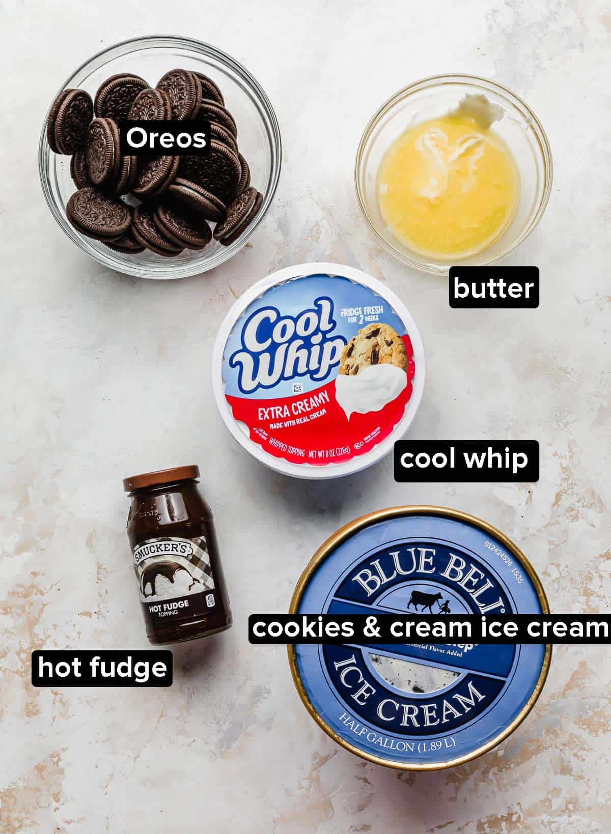 Oreo Ice Cream Cake ingredients in glass bowls: Oreos, butter, oreo ice cream, cool whip, and hot fudge.