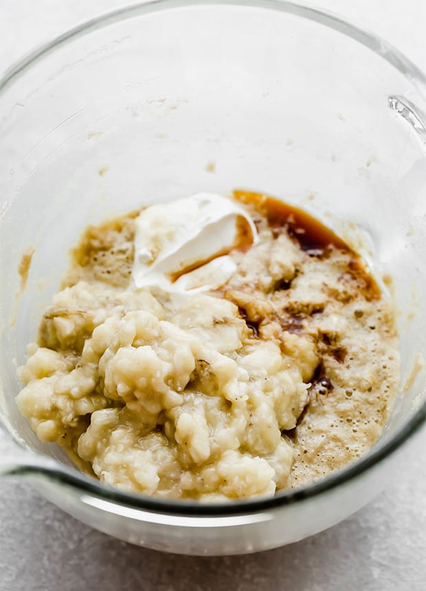 A glass stand mixer bowl with the bread batter and sour cream, mashed bananas, and vanilla extract.