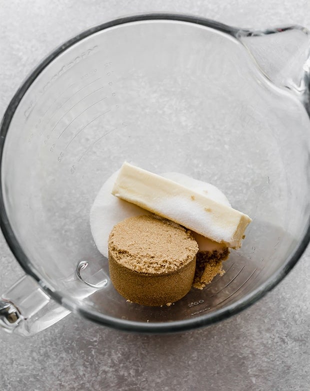 A glass stand mixer bowl with a cube of butter, sugar, and brown sugar in it.