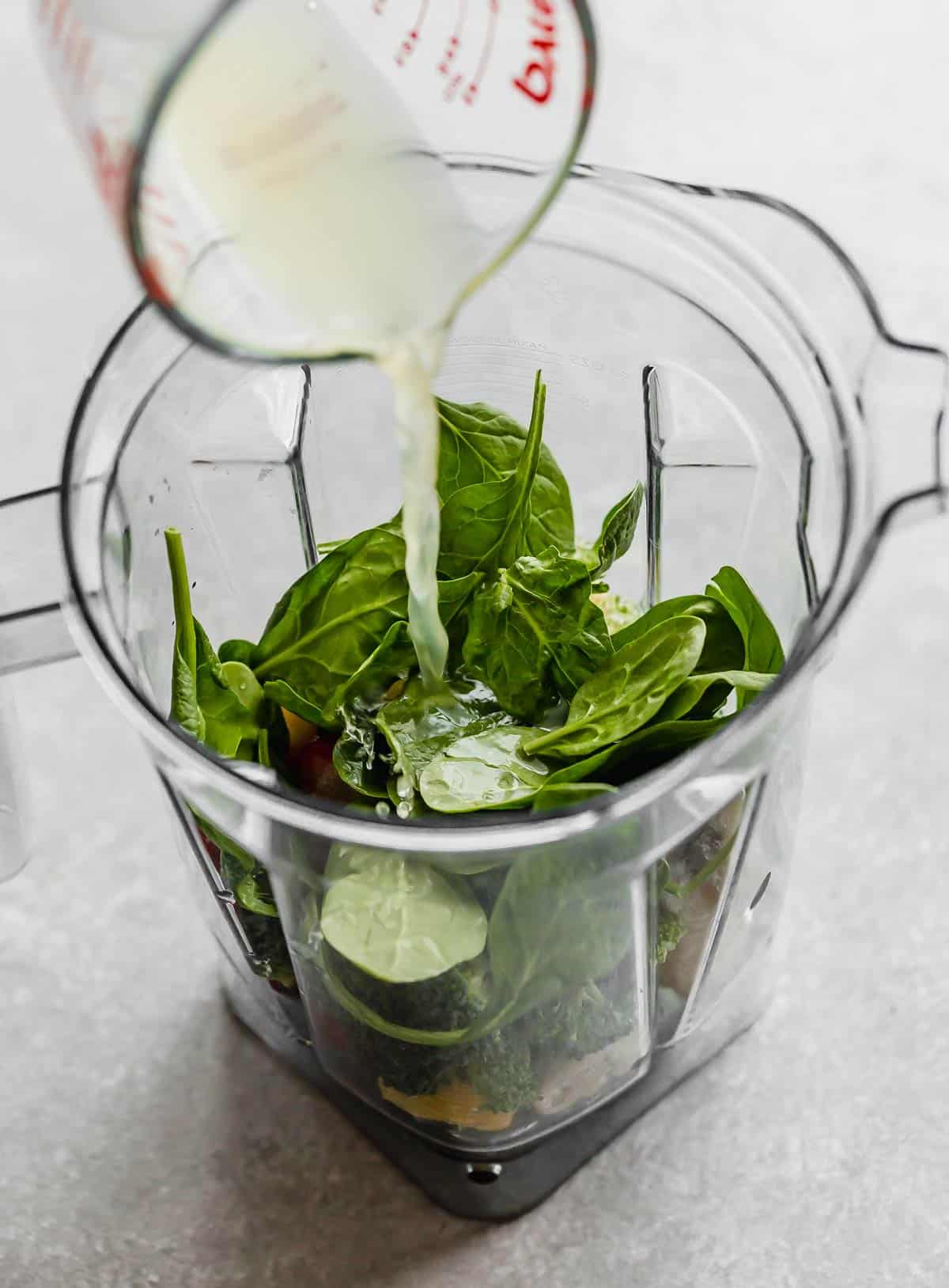 Pineapple juice being poured into a blender that has spinach, broccoli, and fruit in it.