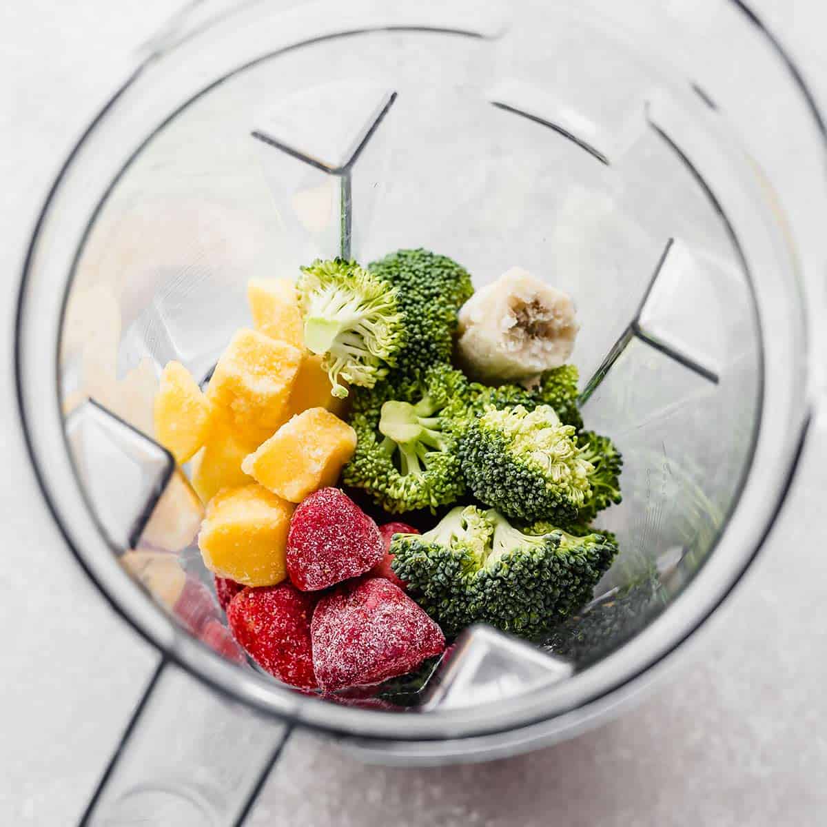 A blender filled with raw broccoli, strawberries, mango chunks, and banana for making a Broccoli Smoothie.