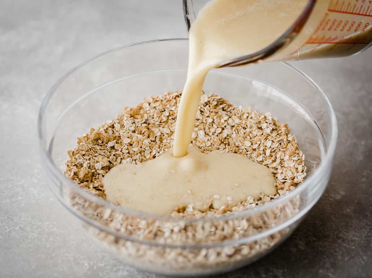 A light yellow colored liquid being poured into a glass bowl full of quick oats.