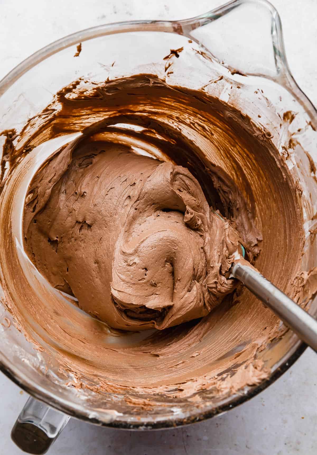 A spatula stirring a thick and creamy chocolate buttercream frosting made with melted chocolate, in a glass bowl.