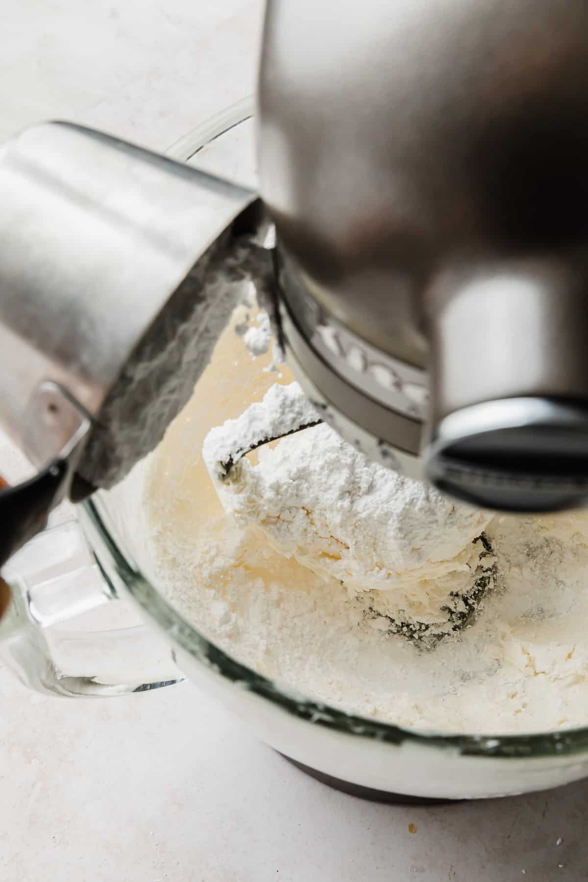 Powdered sugar being poured into a stand mixer bowl that has creamed butter in it, for making Chocolate Buttercream Frosting with melted chocolate.