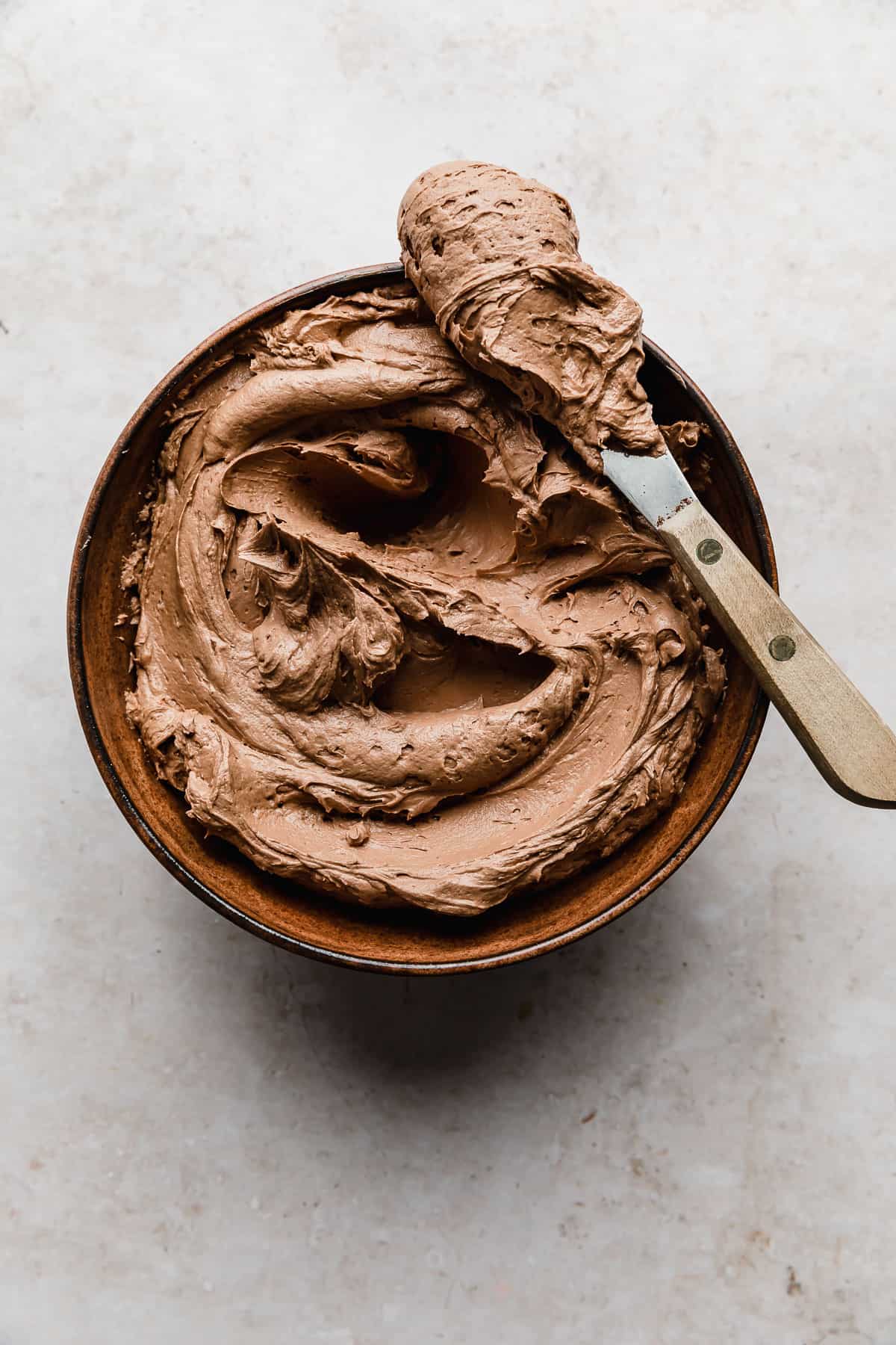 Chocolate Buttercream Frosting Recipe in a brown bowl on a cream textured background.