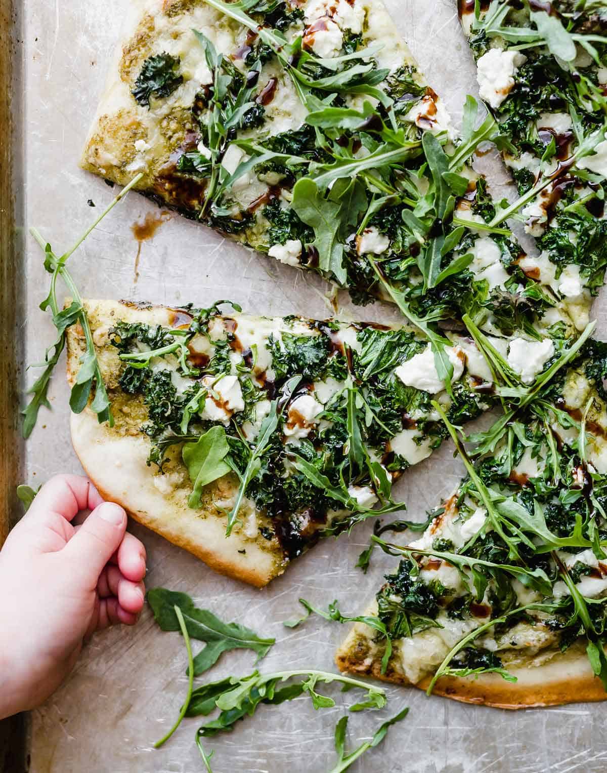 A small child's hand grabbing a slice of homemade kale pizza topped with arugula and balsamic glaze.