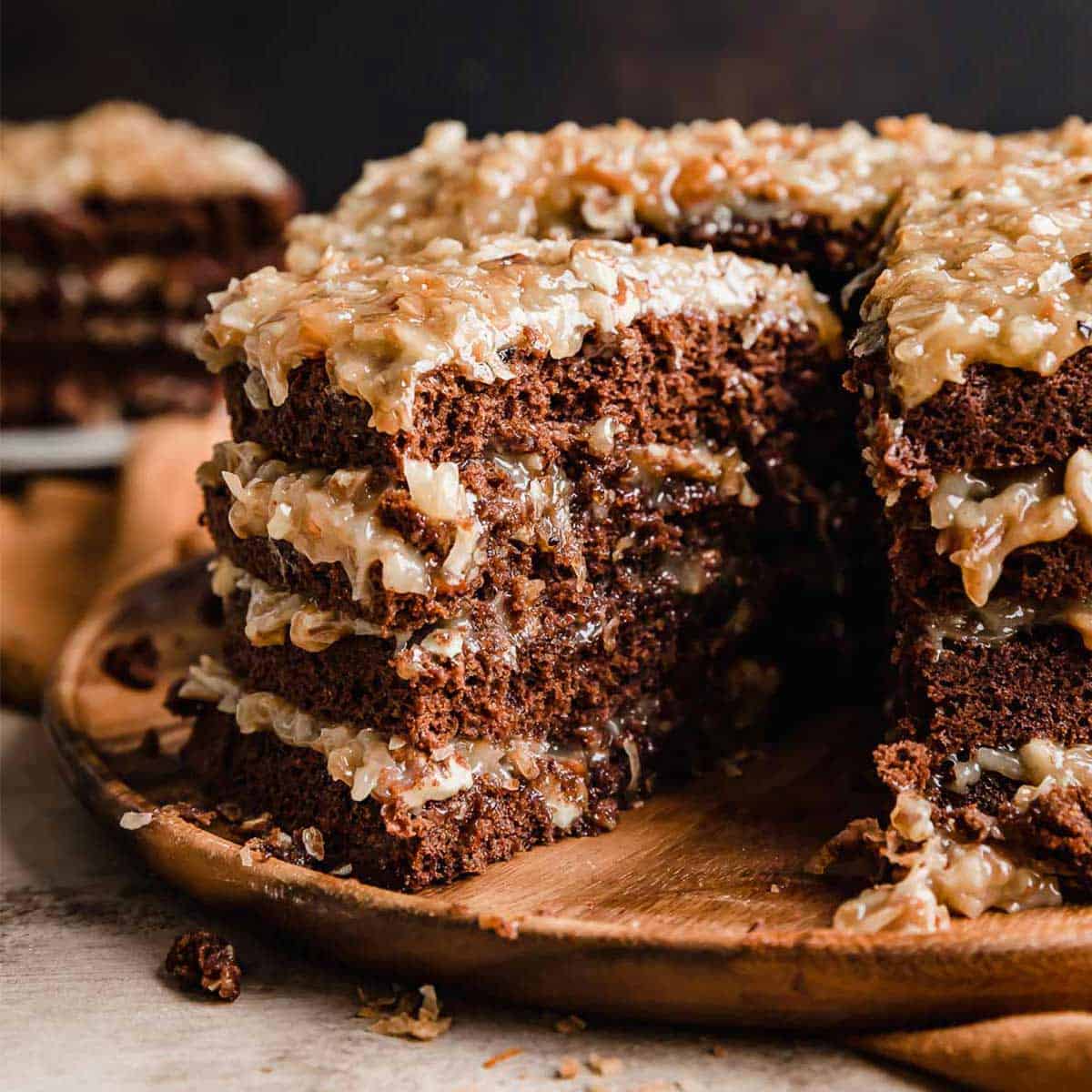 A slice of Traditional German Chocolate Cake taken from a four layer Traditional German Chocolate Cake on a wooden cake plate.