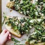A hand grabbing a slice of kale pizza with arugula and goat cheese.