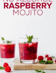 A glass cup on a wooden cutting board full of raspberry mojito mocktail with scattered raspberries and mint leaves near the cup.