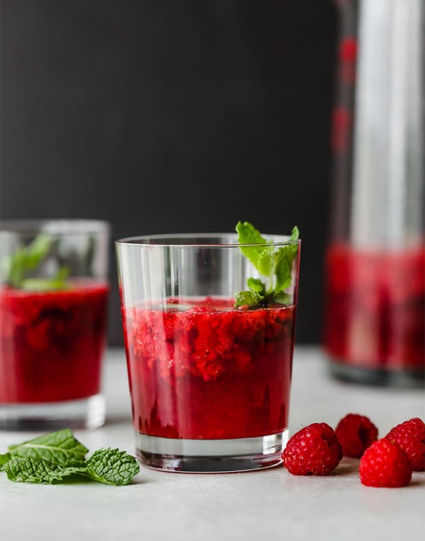 A glass of raspberry mojito with a mint leave in the drink and the pitcher full of the remaining drink in the background.