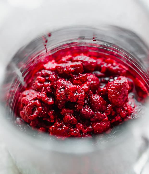 A close up photo of a glass pitcher full of thawed previously frozen raspberries in the pitcher.