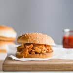 A BBQ chicken sandwich on a wooden board, with hamburger buns stacked in the background.