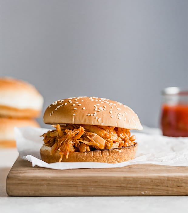 A BBQ chicken sandwich on a wooden board, with hamburger buns stacked in the background.