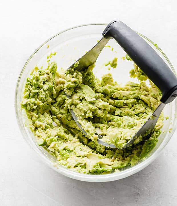 A glass bowl with a pastry cutter mashing avocados.