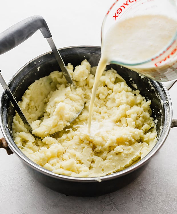 A cream and butter mixture being poured into a pot full of mashed potatoes.