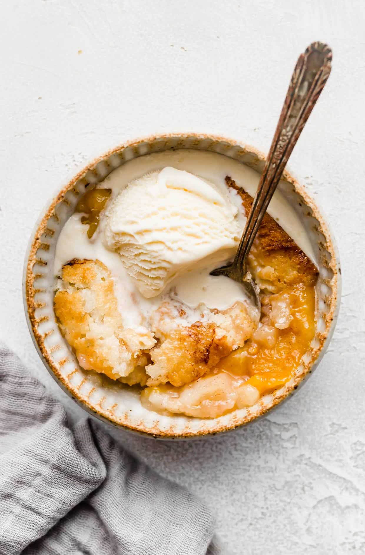 Old Fashioned Peach Cobbler recipe in a tan bowl with melted vanilla ice cream overtop.