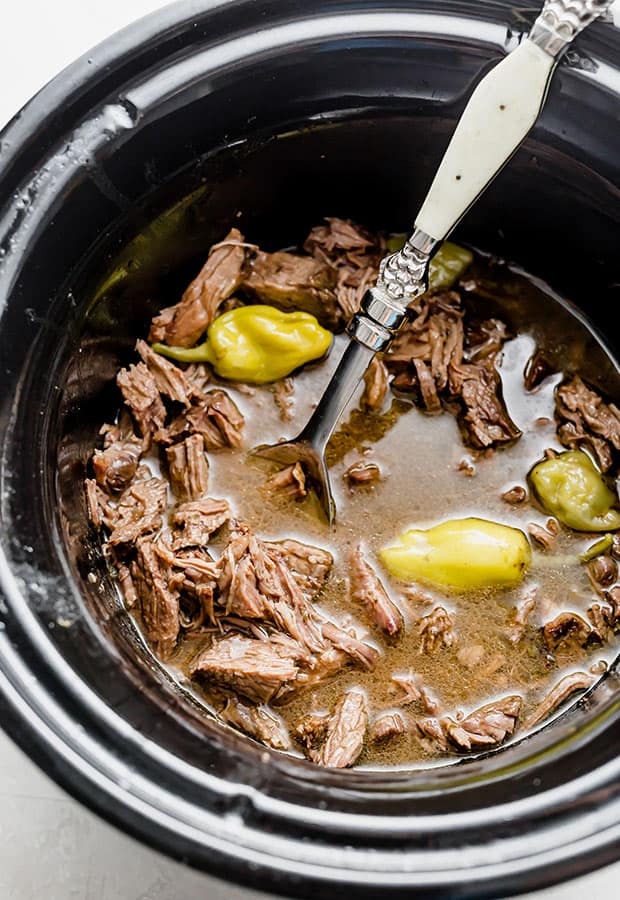 Shredded beef in a slow cooker surrounded by green peppercini peppers.