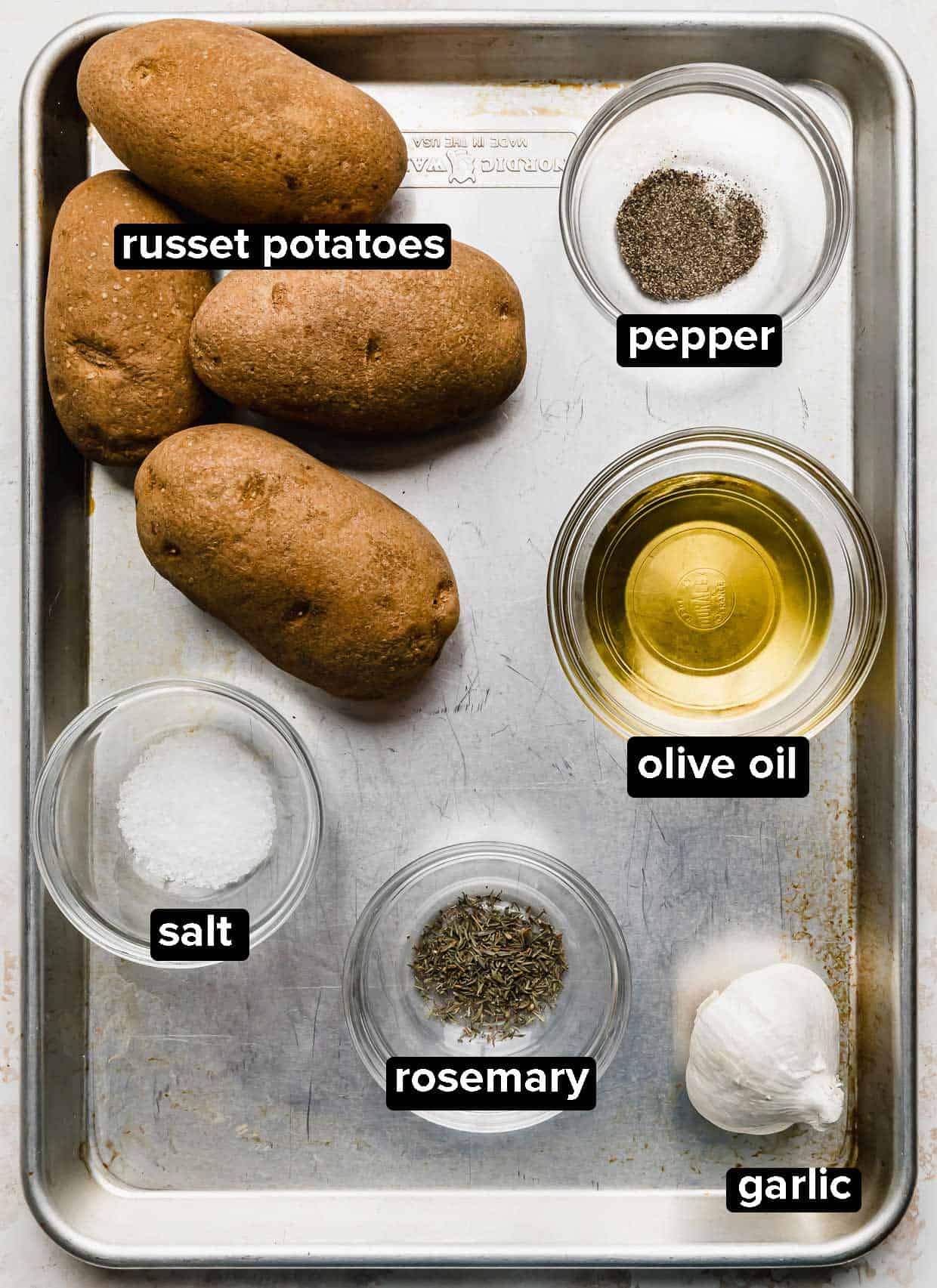 A baking sheet with salt, russet potatoes, garlic, olive oil, garlic, and rosemary on it.