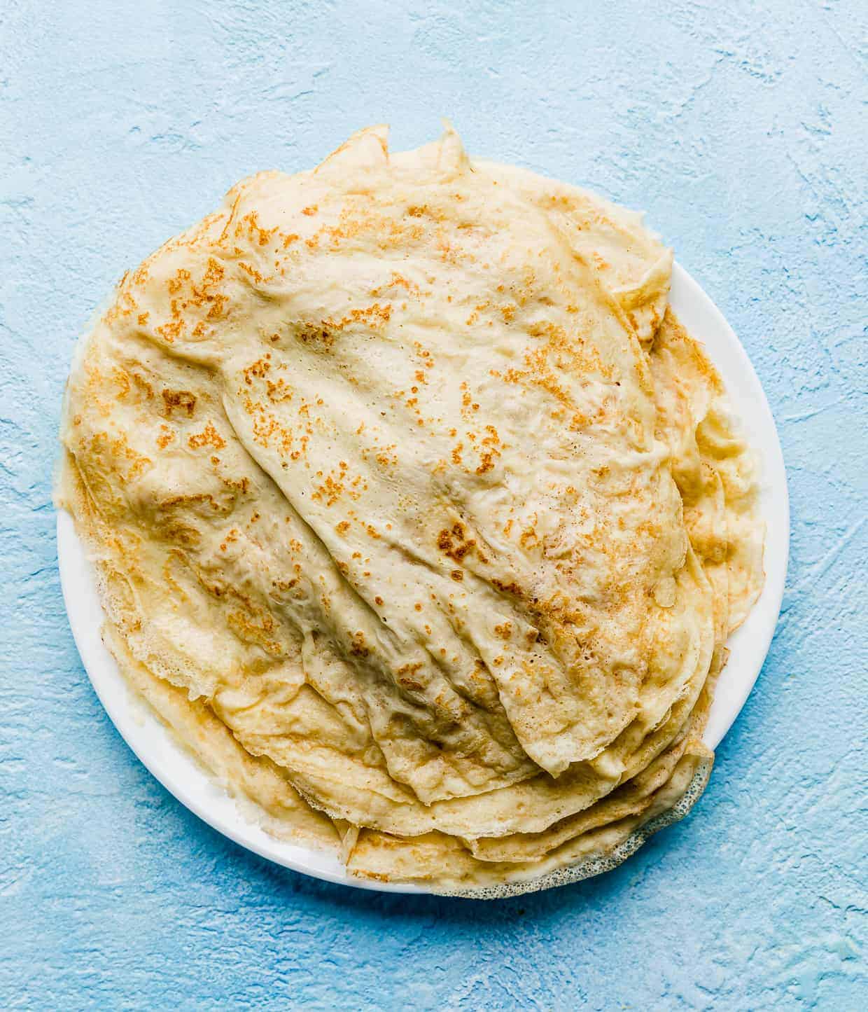 A stack of cooked crepes on a plate on a blue background.