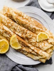 Rolled up English Pancakes on a plate with sliced lemons overtop.