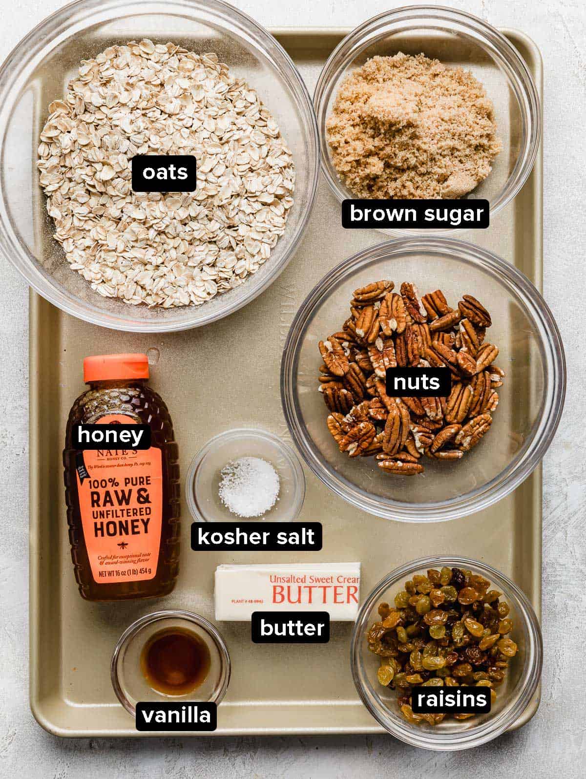 Homemade granola bar ingredients in glass bowls on a bronze colored baking sheet.