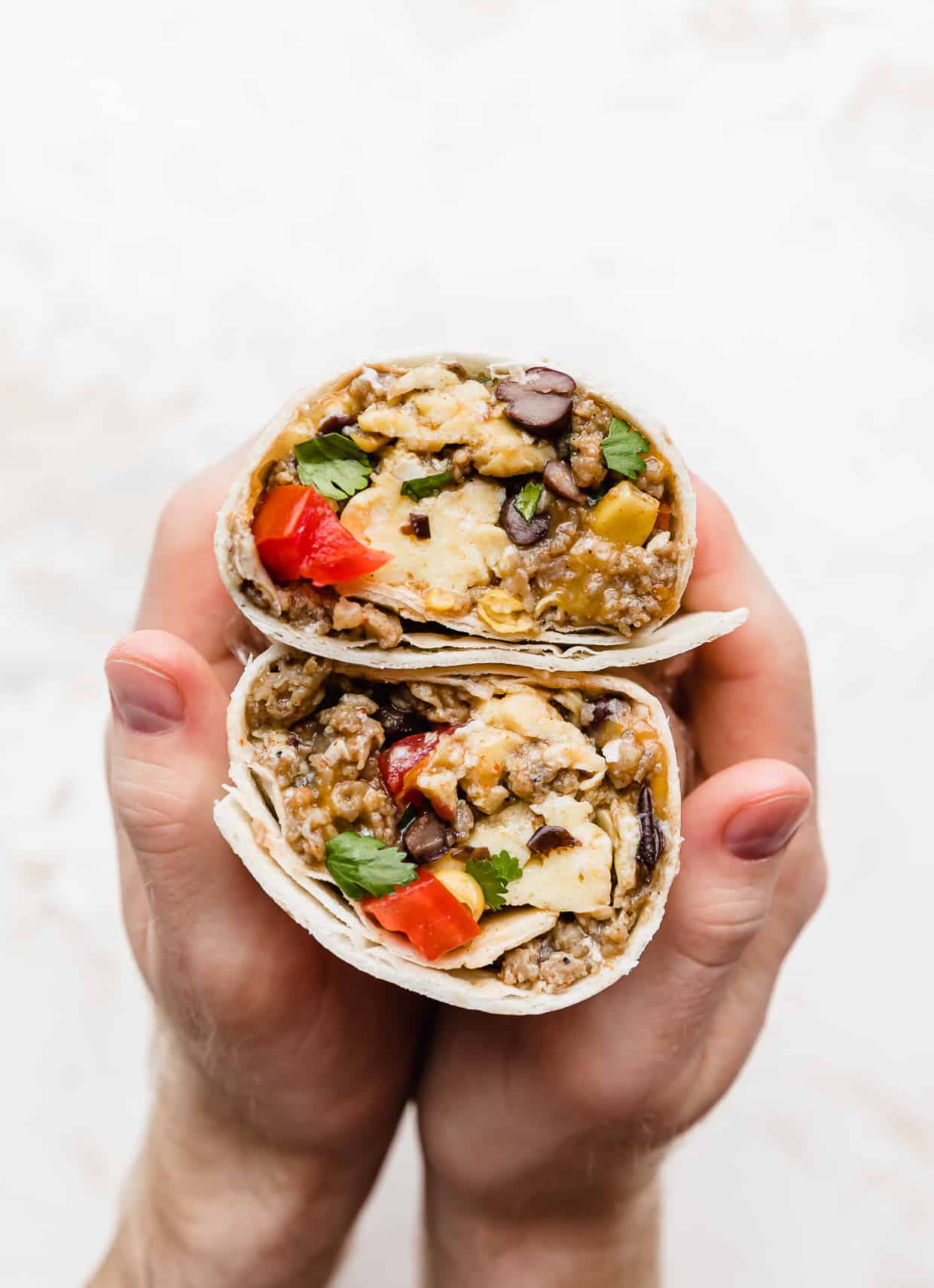 A set of hands holding a Breakfast Burrito with Black Beans and tomatoes that has been cut in half, to show the inside.