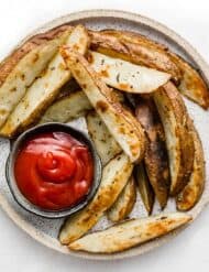 A round white plate with Crispy Baked Potato Wedges on it with a bowl of ketchup, on a white background.
