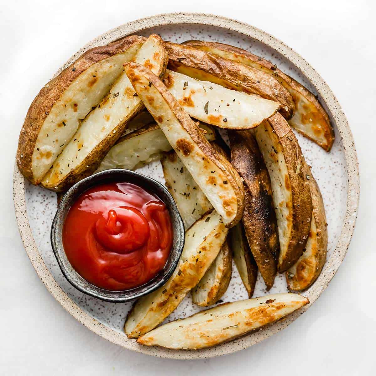 A round white plate with Crispy Baked Potato Wedges on it with a bowl of ketchup, on a white background.