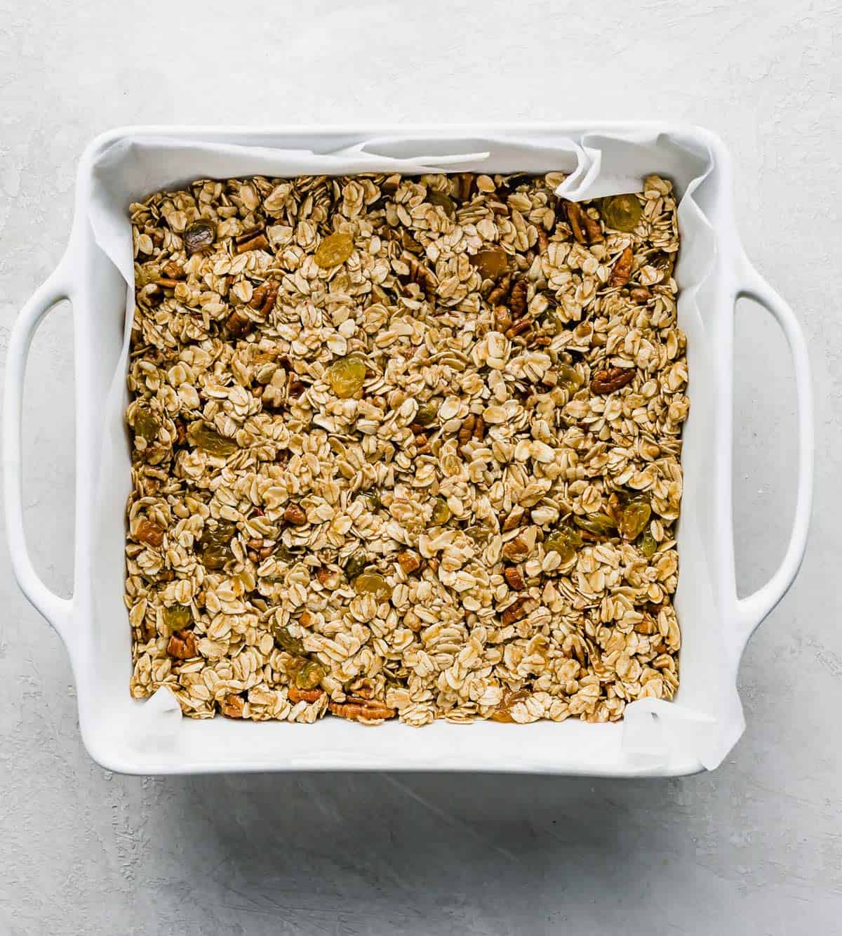 A white square baking dish filled with an Oatmeal Raisin Granola Bar mixture, o na white background.