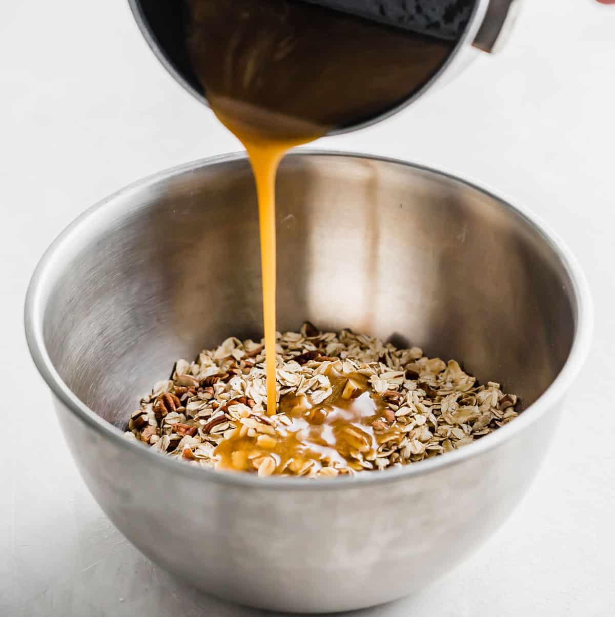 A brown liquid being poured from a small pot into a metal bowl filled with Oatmeal Raisin Granola Bar mixture.