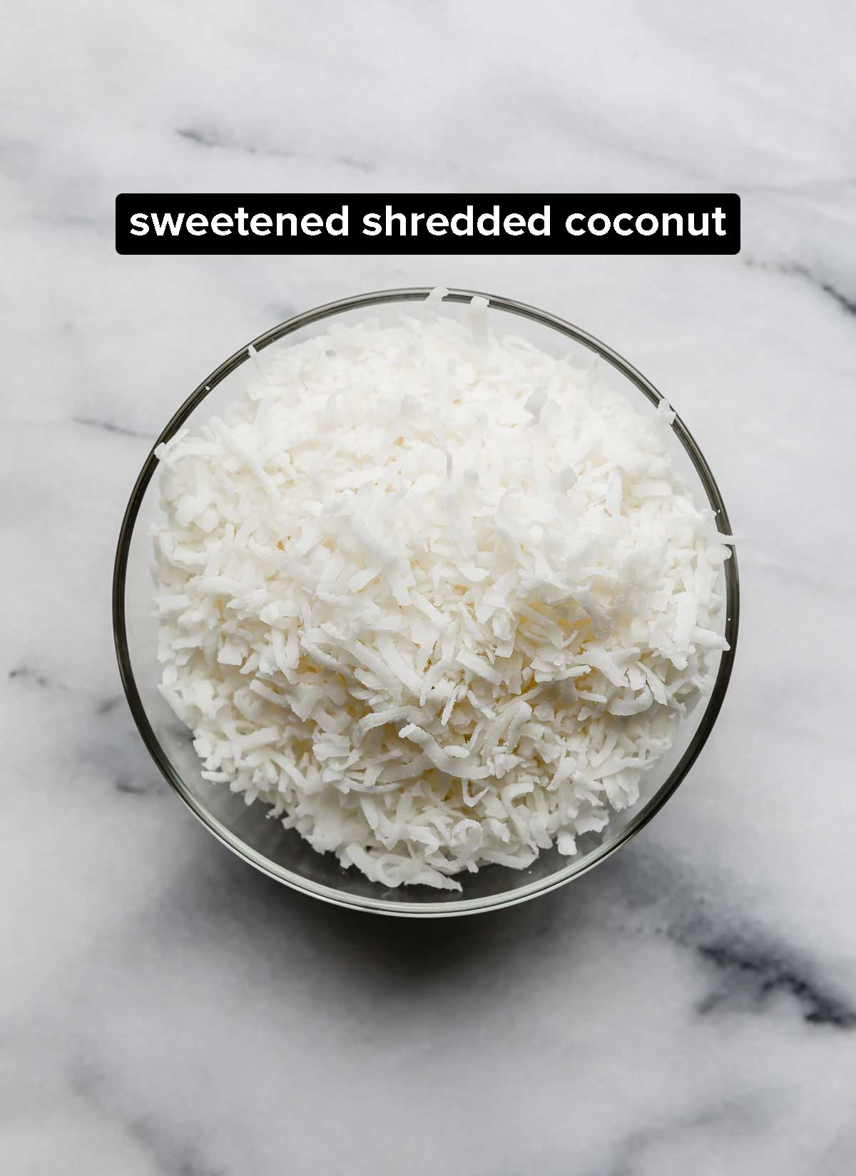 Sweetened shredded coconut in a glass bowl on a white marble background.