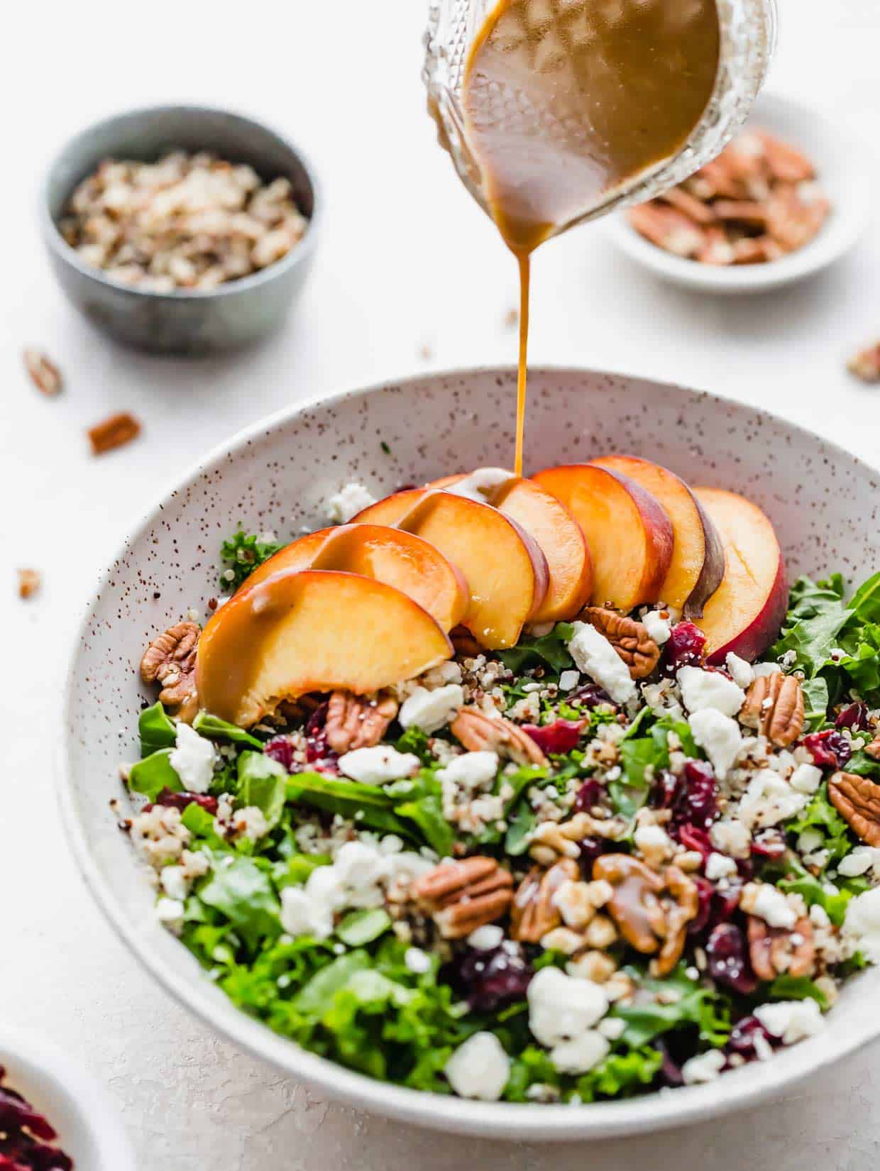 Balsamic dressing being poured overtop a kale and quinoa salad with nuts, raisins, feta cheese and sliced peaches.