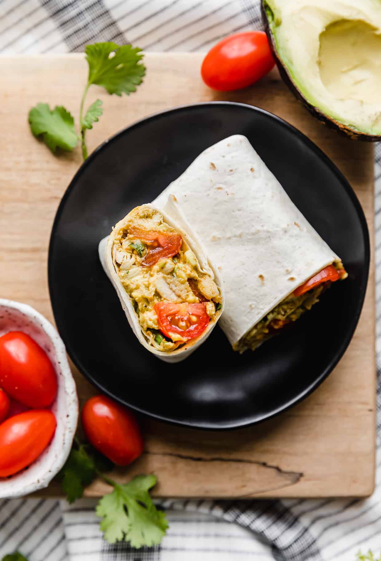 An Avocado Chicken Wrap on a black plate with grape tomatoes and sliced avocado surrounding it.