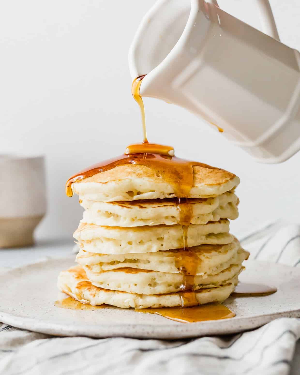 A stack of Homemade Buttermilk Pancakes on a white plate with maple syrup being poured overtop of the stack.