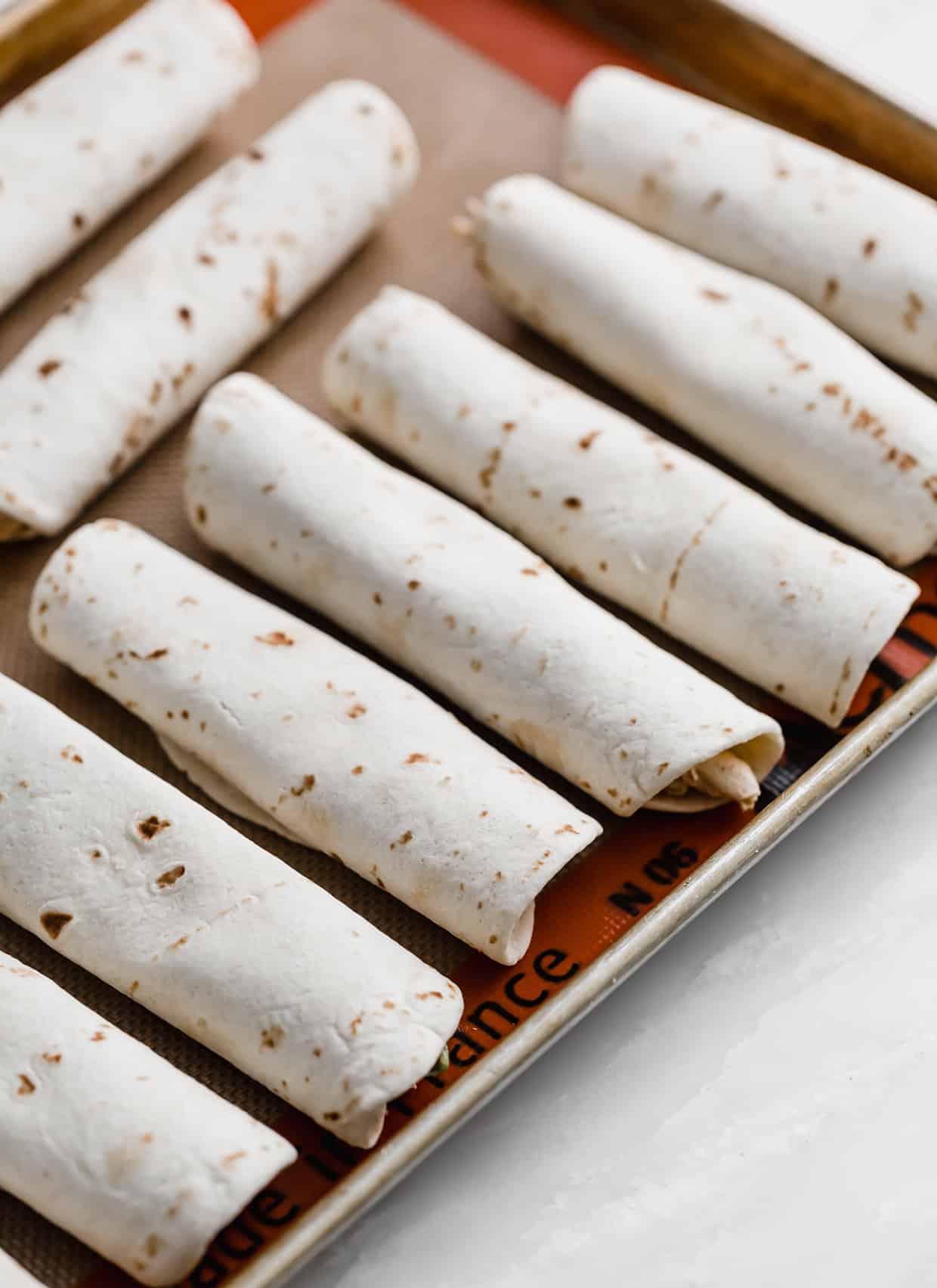 Cream cheese and chicken filled flour tortillas lined up on a baking sheet.
