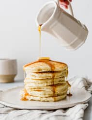 Maple syrup being poured over a stack of fluffy buttermilk pancakes on a white plate.