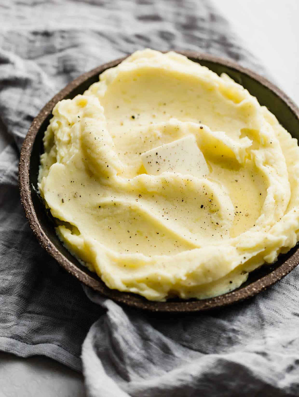 A slice of butter melting into the top of homemade mashed potatoes on a black plate.